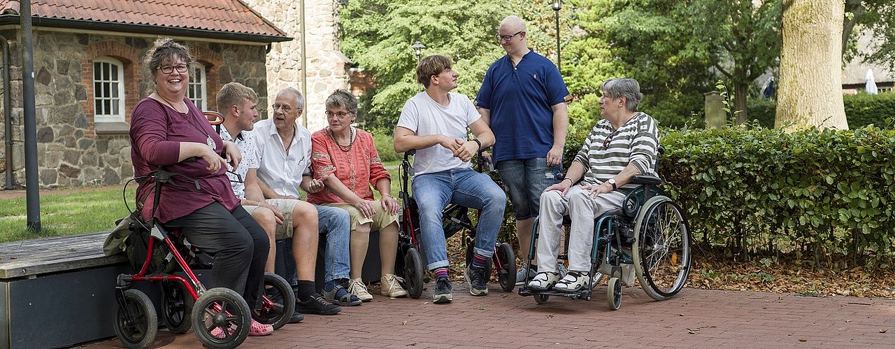 Auf dem Foto zu sehen ist eine bunt gemischte Gruppe jüngerer und älterer Menschen mit und ohne Behinderung, die sich draußen trifft 