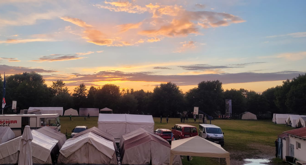 Das Bild ist eine Aufnahme des Zeltplatzes bei Sonnenuntergang. Zu sehen ist der Abendhimmel über den weißen Gruppenzelten