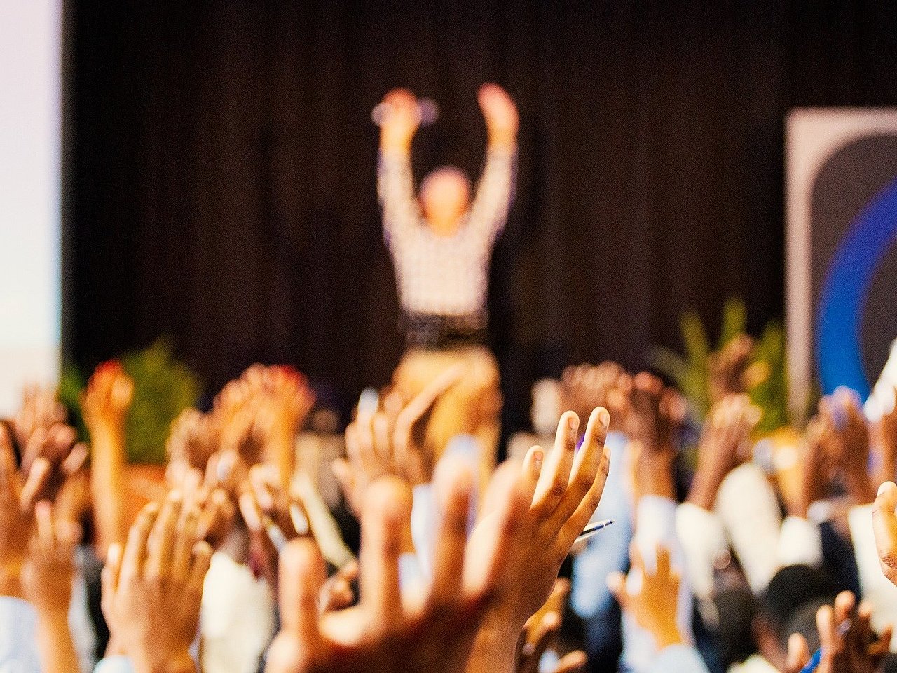 Das Foto zeigt verschwommen einen Menschen vor einem Publikum, das die Hände in die Höhe reckt